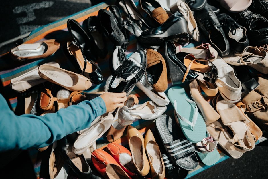 A person wearing a blue long-sleeve shirt is selecting and holding a shiny silver shoe from a large collection of various footwear arranged on a striped, multicolored surface. The assortment includes 