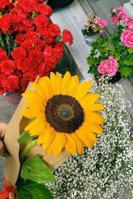 A vibrant sunflower with bright yellow petals and a large dark brown center, wrapped in light brown kraft paper with green leaves visible at the base, held by a person in front of a display of fresh f