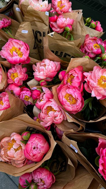 An arrangement of fresh pink peonies with lush, ruffled petals and bright yellow centers, wrapped in light brown kraft paper and tied with natural twine. The flowers are set against a dark background,
