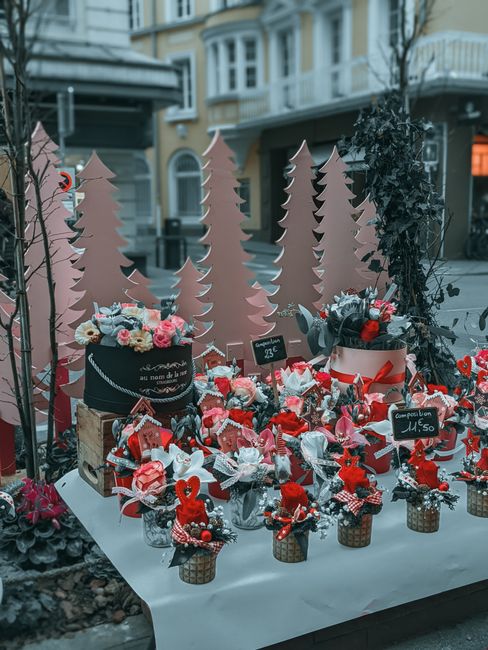 A floral display at an outdoor market stall featuring a variety of colorful flower arrangements and gift items. The arrangement includes bouquets of pink, red, and white flowers, with some decorated w