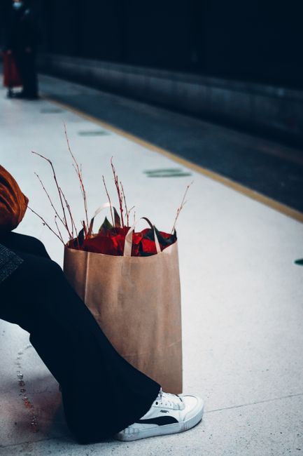A person sitting on the floor at Brockley Station platform, holding a large brown paper bag filled with a floral gift. Inside the bag, vibrant red flowers with green foliage are visible, accented by w