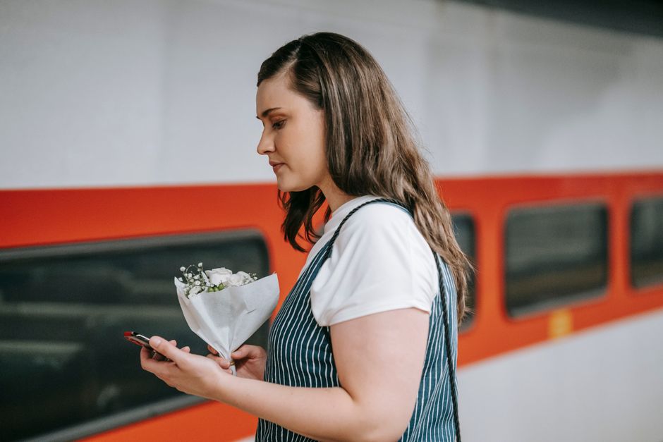 A woman with long brown hair, wearing a white t-shirt and a blue striped apron, is holding a small bouquet of fresh white flowers wrapped in white paper. She is standing in a station with a blurred re