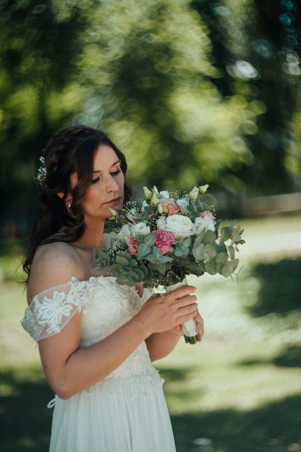 A bride with dark, wavy hair adorned with small floral accessories holds a lush bouquet of fresh flowers outdoors, near Hilly Fields. The bouquet features a mix of soft pink roses, white peonies, euca